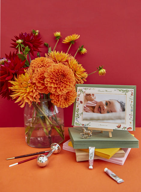 Image of a Clementina Sketchbook printed birth announcement propped up against a vase of orange flowers, surrounded by different instruments and an orange background.