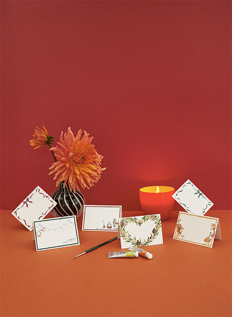 Image of different Clementina Sketchbook printed place cards, propped against a candle and a vase of orange flowers, which are themselves placed in front of a red background. 