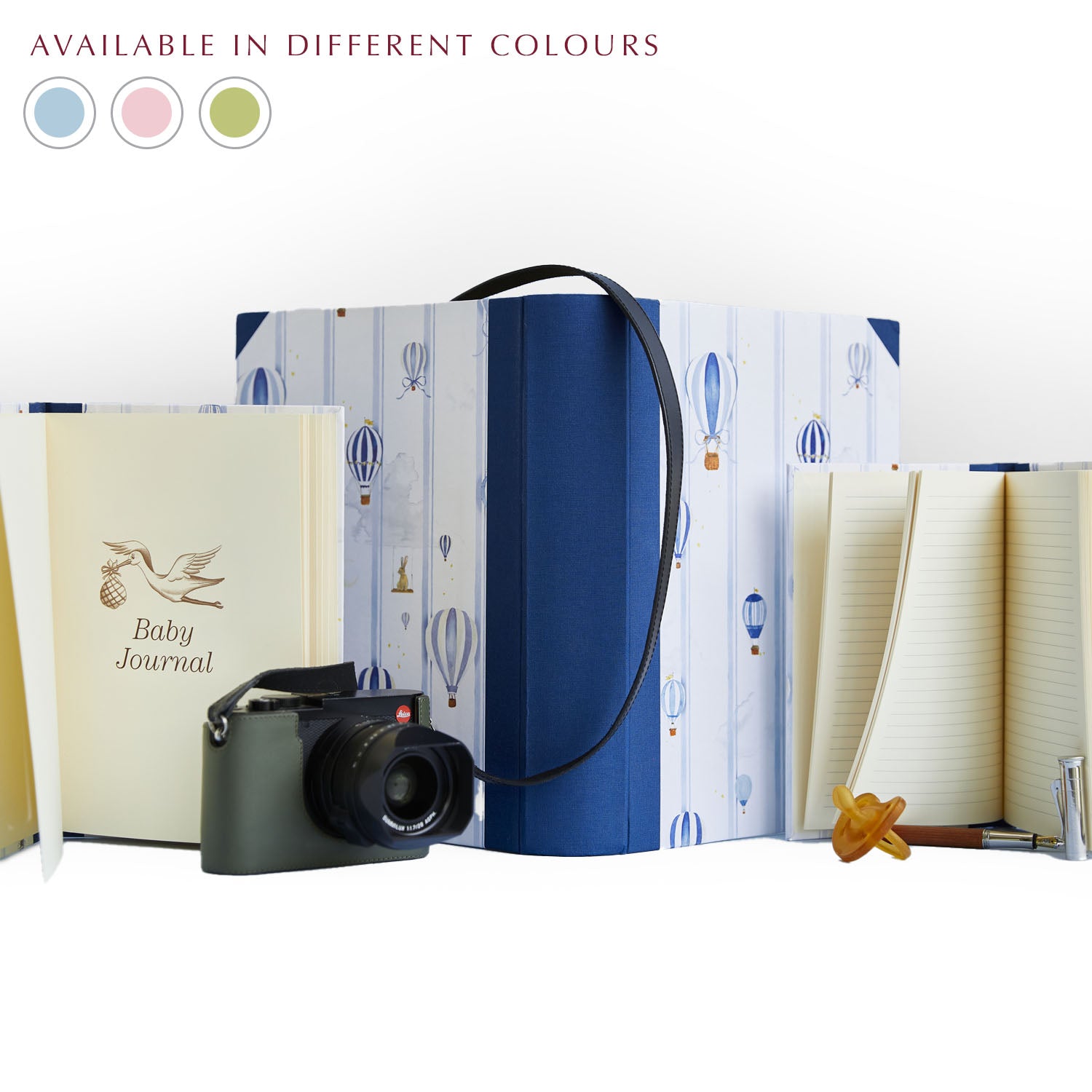 Image of Clementina Sketchbook's hot air balloon baby book bundle in blue against a green background. In the corner of the image is a colour wheel showing the different colours the bundle is available: blue, pink and green. A camera, fountain pen and pacifier lie nearby. 