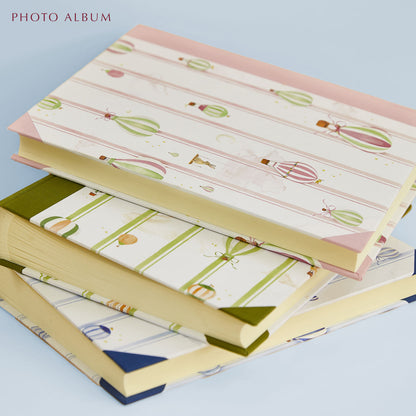 Close-up of the three different baby photo albums in pink, green and blue stacked on top of one another against a green background. Each features hand-painted watercolour designs.