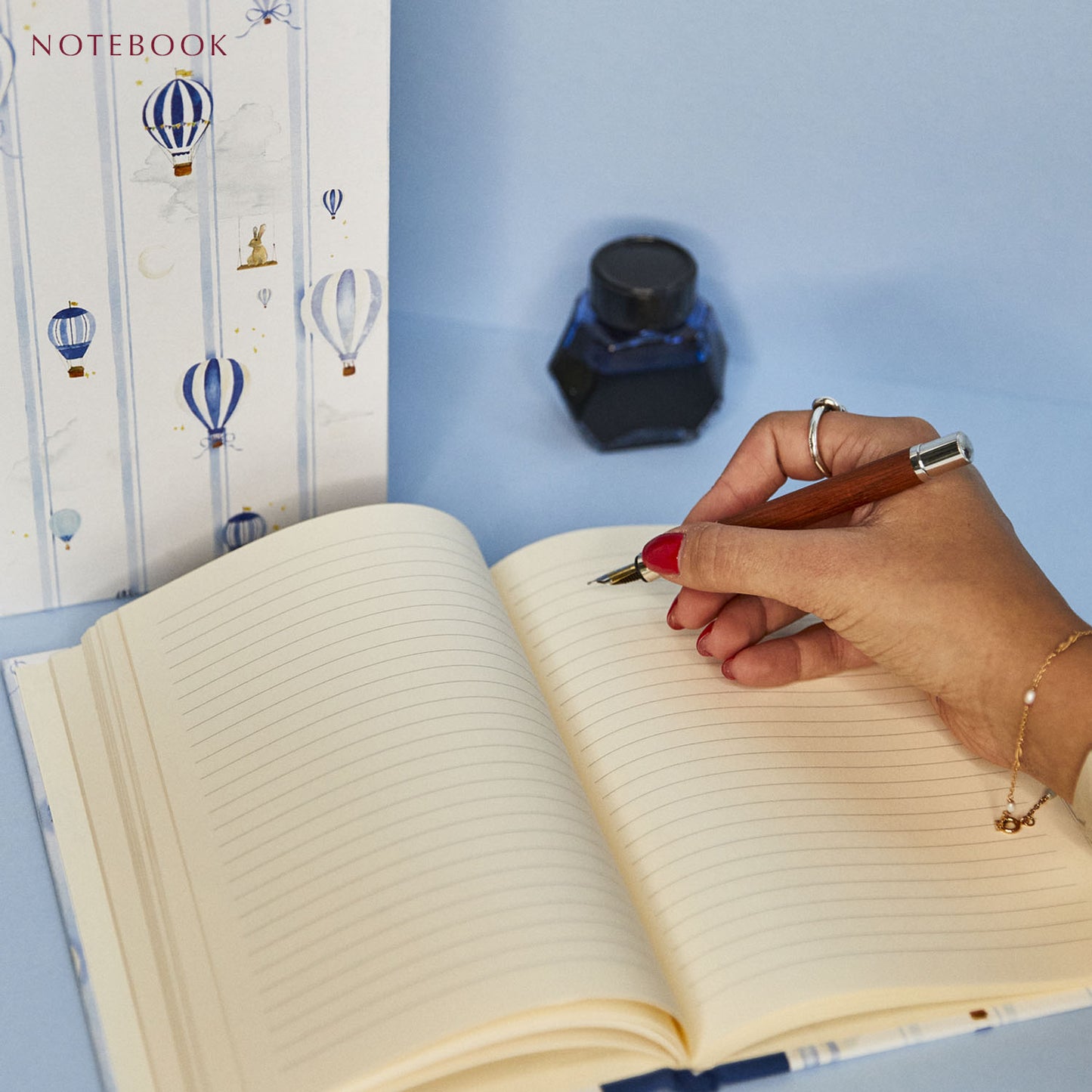 Image of a parent writing in the baby book bundle's notebook using a fountain pen. A pot of ink lies nearby.