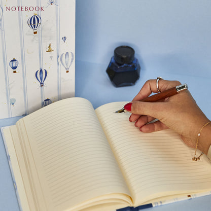 Image of a parent writing in the baby book bundle's notebook using a fountain pen. A pot of ink lies nearby.