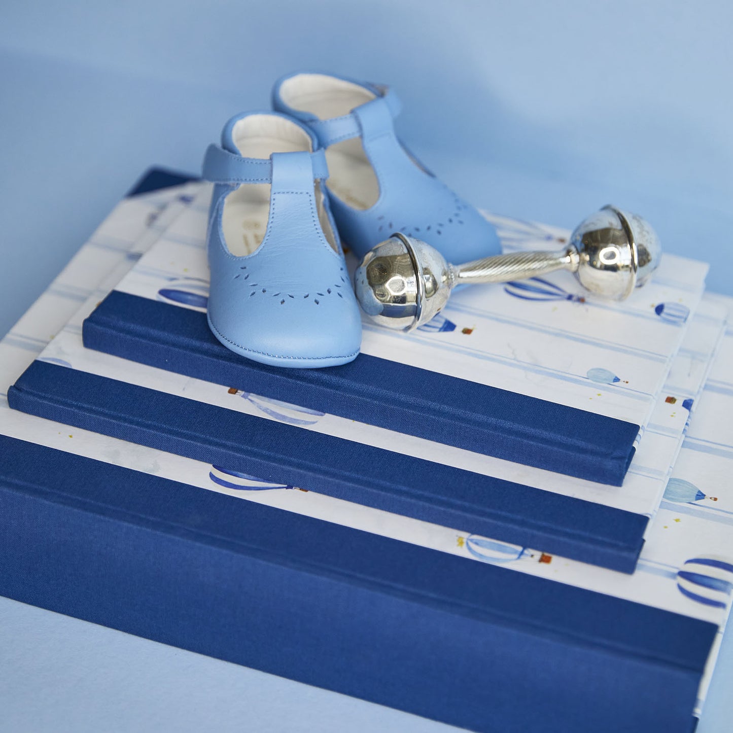 Image of the hot air balloon baby book bundle in blue stacked on top of one another with a child's rattle and pair of shoes resting on top. 